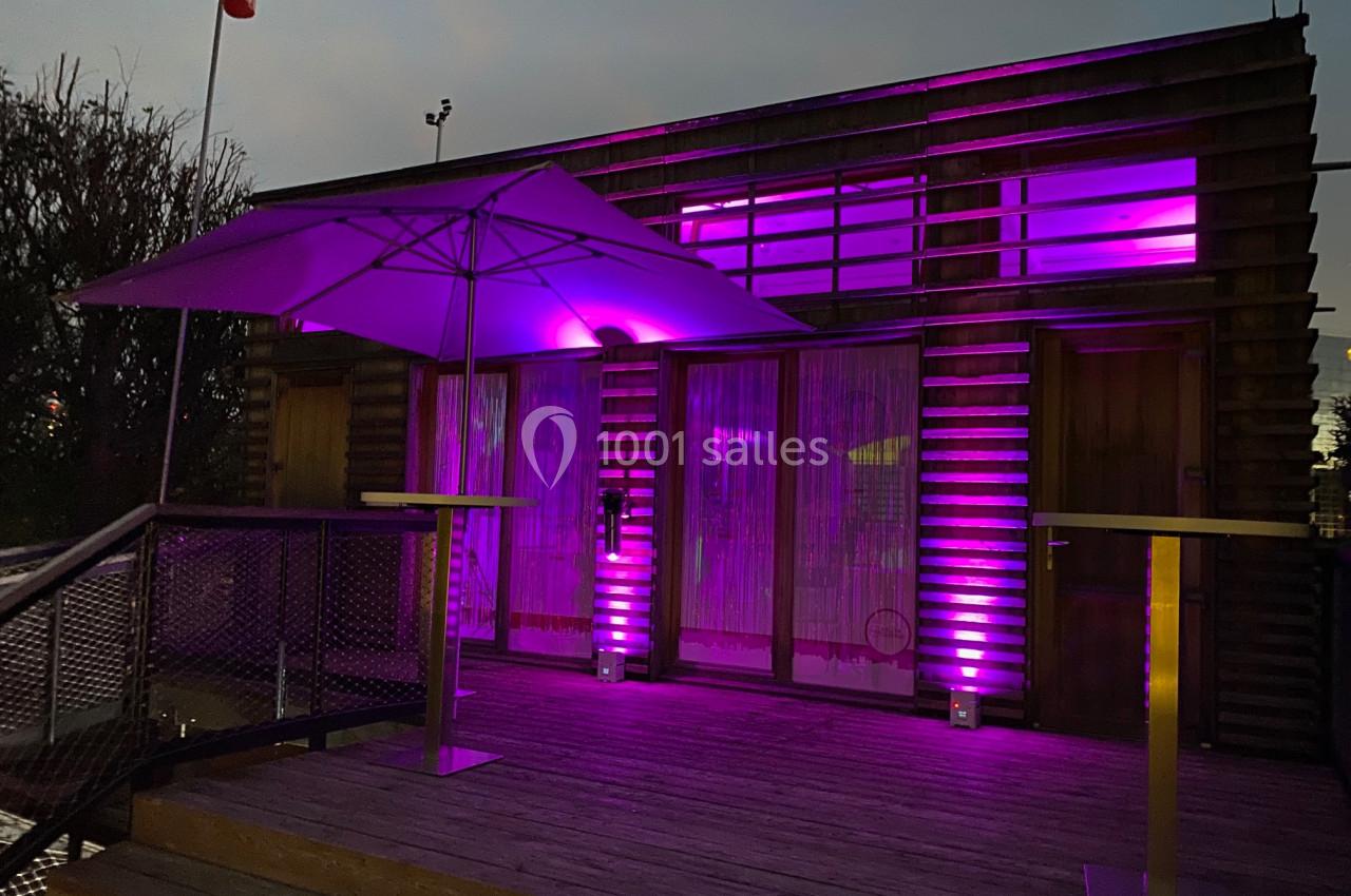 Façade en bois éclairée par des lumières violettes, avec une terrasse en bois et un grand parasol.