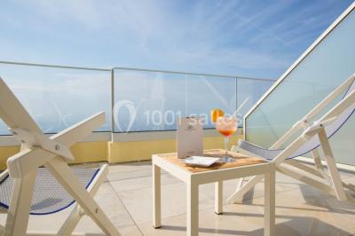 Terrasse ensoleillée avec tables hautes dressées, vue sur la mer et des arbres en arrière-plan.