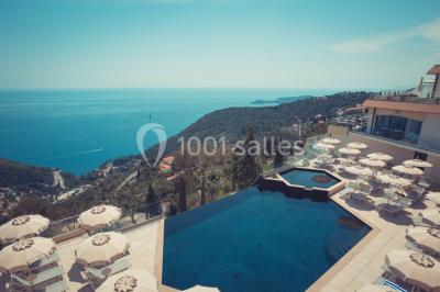 Terrasse ensoleillée avec tables hautes dressées, vue sur la mer et des arbres en arrière-plan.