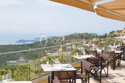 Terrasse ensoleillée avec tables hautes dressées, vue sur la mer et des arbres en arrière-plan.