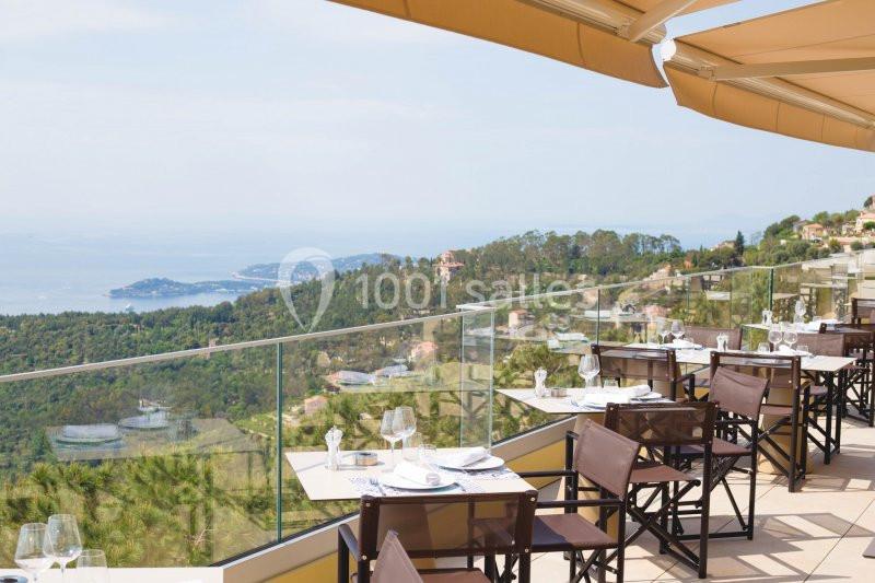Terrasse d'un restaurant avec tables dressées, offrant une vue panoramique sur des collines verdoyantes et la mer au loin.