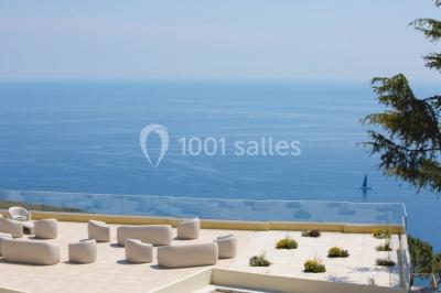 Terrasse ensoleillée avec tables hautes dressées, vue sur la mer et des arbres en arrière-plan.