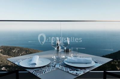 Terrasse ensoleillée avec tables hautes dressées, vue sur la mer et des arbres en arrière-plan.