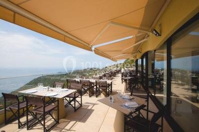 Terrasse ensoleillée avec tables hautes dressées, vue sur la mer et des arbres en arrière-plan.