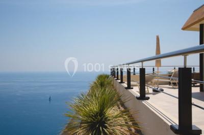 Terrasse ensoleillée avec tables hautes dressées, vue sur la mer et des arbres en arrière-plan.