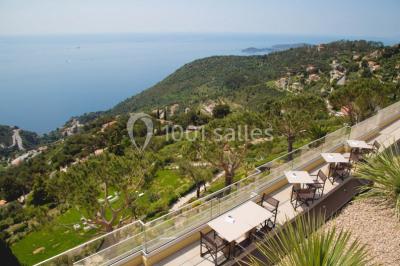 Terrasse ensoleillée avec tables hautes dressées, vue sur la mer et des arbres en arrière-plan.