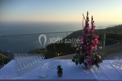 Terrasse ensoleillée avec tables hautes dressées, vue sur la mer et des arbres en arrière-plan.