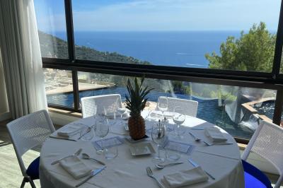 Terrasse ensoleillée avec tables hautes dressées, vue sur la mer et des arbres en arrière-plan.