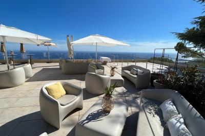 Terrasse ensoleillée avec tables hautes dressées, vue sur la mer et des arbres en arrière-plan.