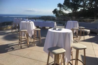Terrasse ensoleillée avec tables hautes dressées, vue sur la mer et des arbres en arrière-plan.