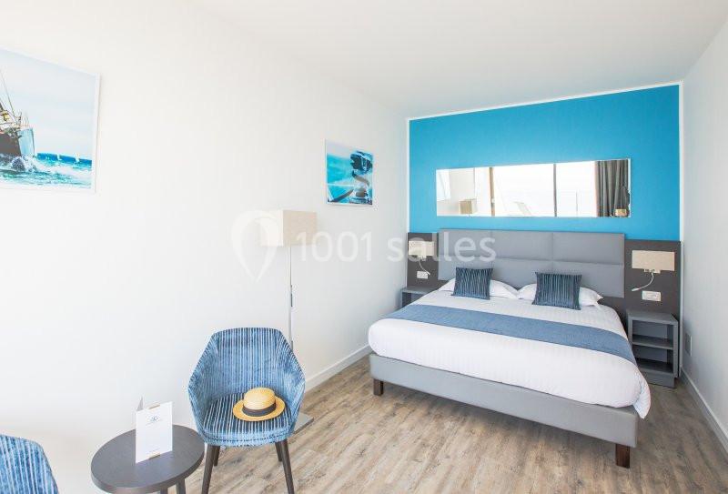 Chambre lumineuse avec un lit double, mur bleu, fauteuil, table avec un chapeau et décorations marines.