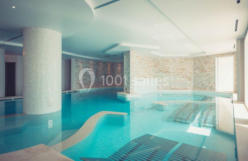 Piscine intérieure moderne avec colonnes, murs en pierre et lumière naturelle entrant par une grande baie vitrée.