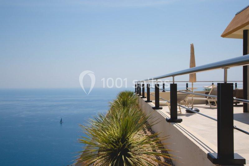 Vue d'une terrasse en bord de mer avec garde-corps en métal, parasol et plantes, surplombant l'eau calme.