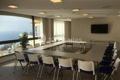 Salle de réunion lumineuse avec tables disposées en U, chaises blanches, bouteilles d'eau et écran de projection.