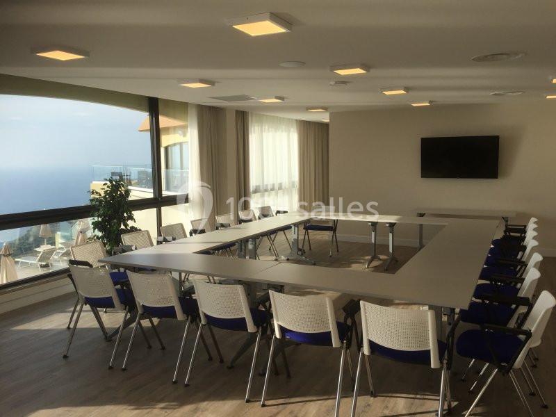 Salle de réunion lumineuse avec grandes baies vitrées, tables en U, chaises blanches et écran mural.