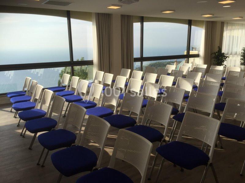 Salle de conférence lumineuse avec des rangées de chaises blanches et bleues, offrant une vue panoramique sur la mer.