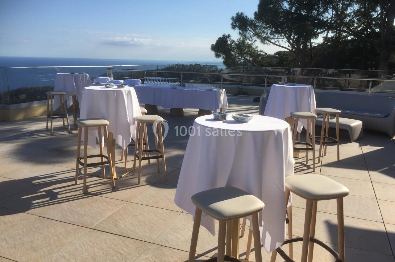 Terrasse ensoleillée avec tables hautes dressées, vue sur la mer et des arbres en arrière-plan.