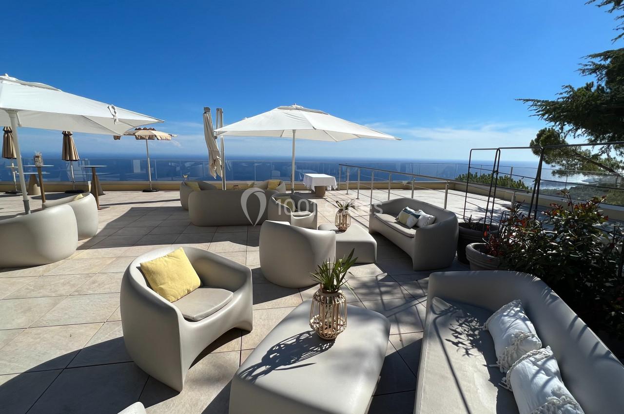 Terrasse ensoleillée avec mobilier moderne, parasols blancs et vue panoramique sur la mer et le ciel bleu.