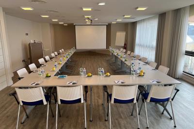 Salle de réunion lumineuse avec tables disposées en U, chaises blanches, bouteilles d'eau et écran de projection.