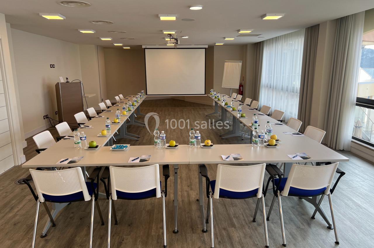 Salle de réunion lumineuse avec tables disposées en U, chaises blanches, bouteilles d'eau et écran de projection.