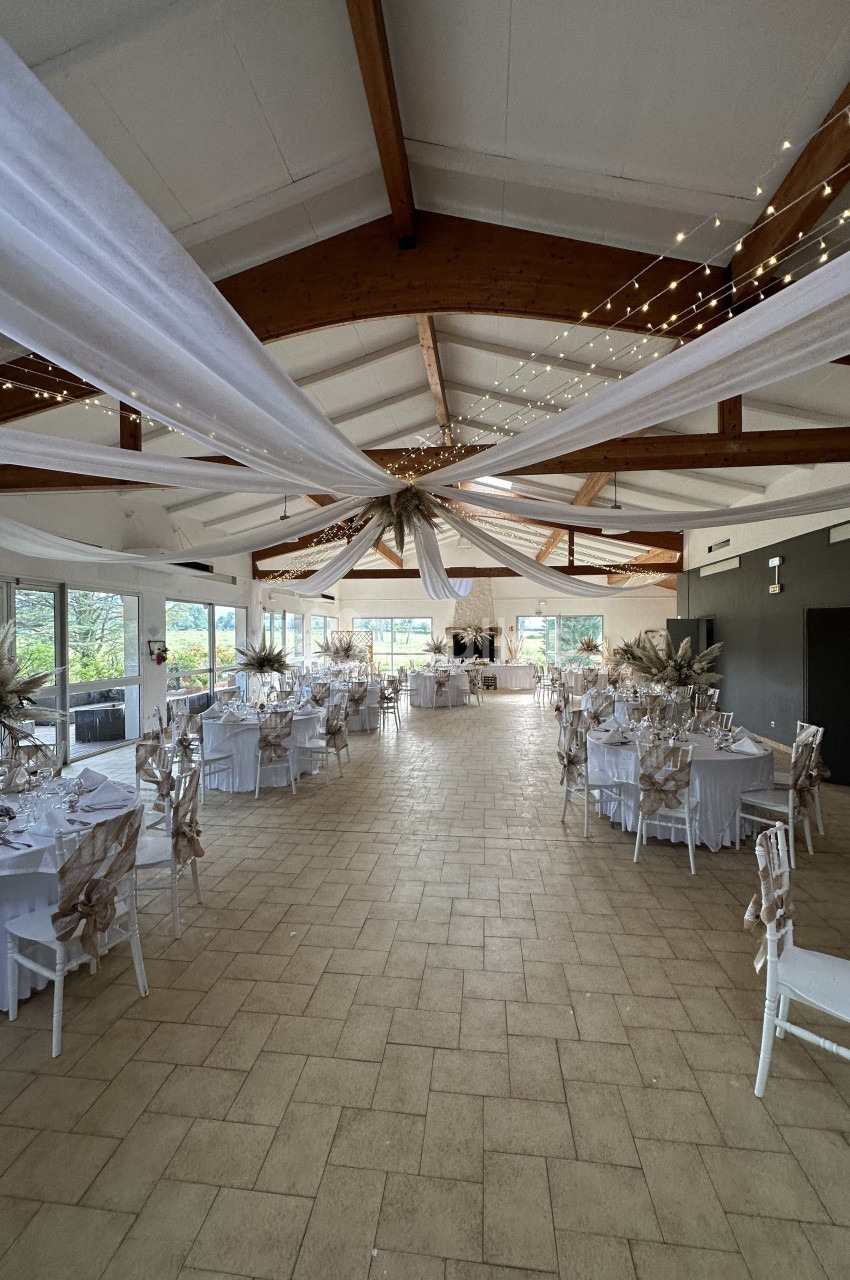 Salle de réception décorée avec des tables rondes, nappes blanches, chaises ornées et voilages suspendus au plafond.