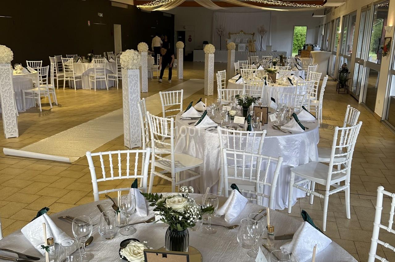 Salle de réception décorée pour un mariage, avec tables rondes dressées, nappes blanches et guirlandes lumineuses suspendues.