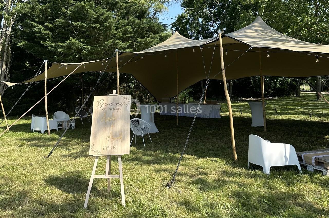 Tente beige montée sur une pelouse, avec des tables et chaises blanches, et un panneau de bienvenue au premier plan.