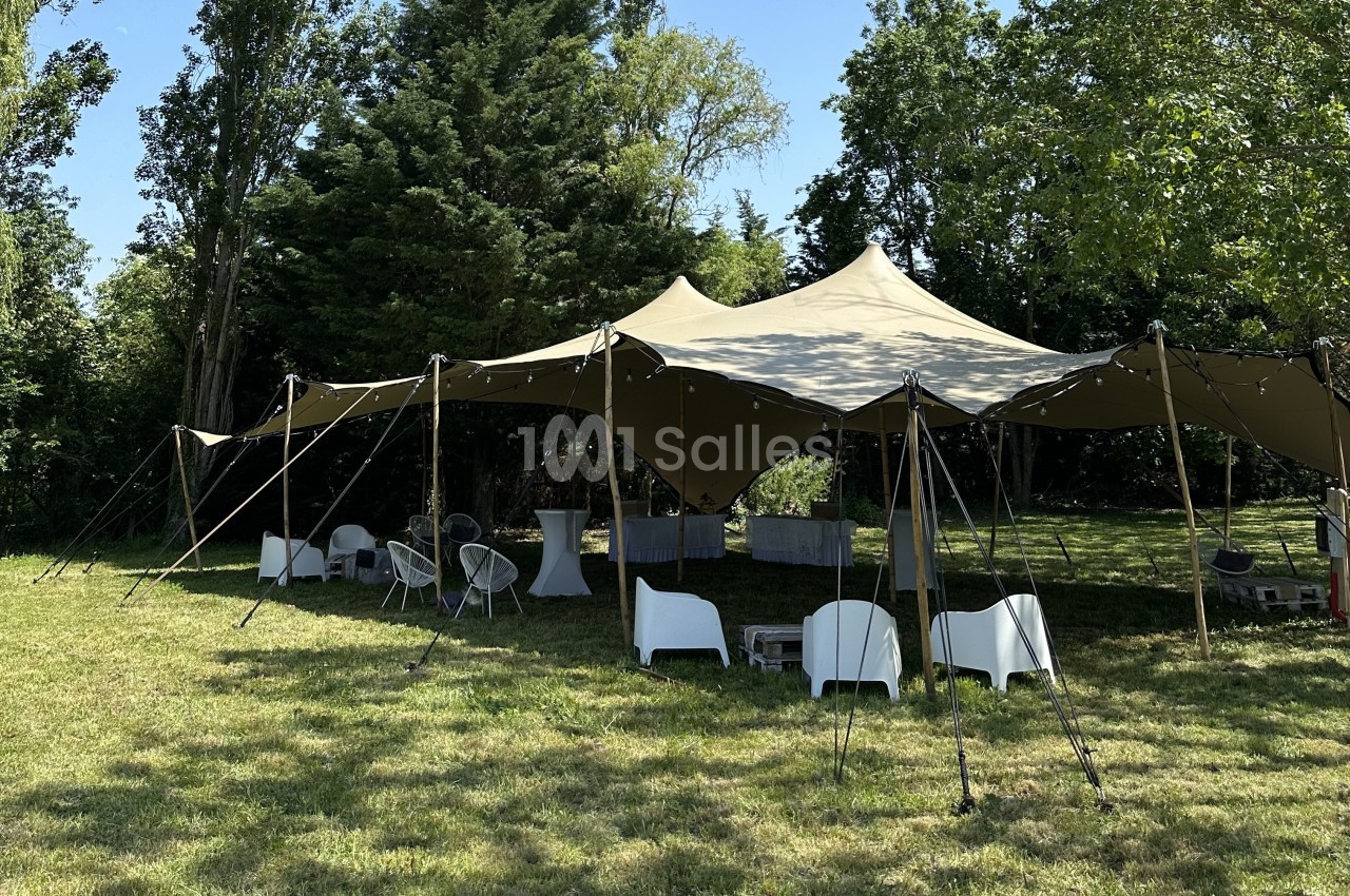 Tente beige installée sur une pelouse, entourée de chaises blanches et de tables, dans un cadre arboré.