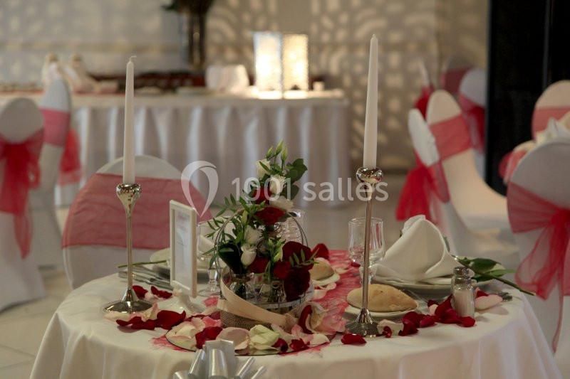 Table décorée pour un événement avec nappes blanches, roses rouges, bougies et chaises ornées de rubans rouges.