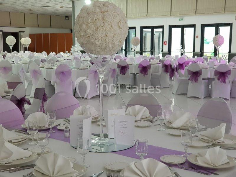 Salle de réception décorée pour un événement, avec tables dressées, nappes blanches et chaises ornées de nœuds violets.