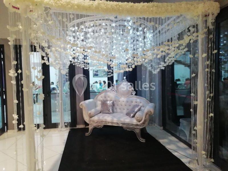 Canapé argenté sous une arche décorée de fleurs blanches et de guirlandes lumineuses dans un espace intérieur.