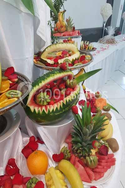 Présentoir de fruits sculptés avec pastèques, ananas, fraises et autres fruits, décoré de pétales rouges.