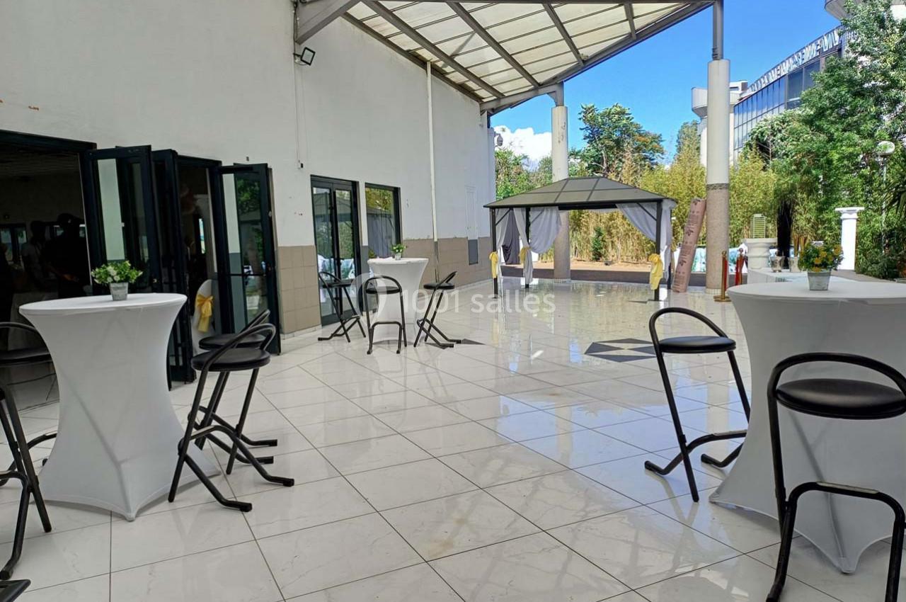 Terrasse couverte avec sol en carrelage blanc, tables hautes, chaises noires et tonnelle dans un espace extérieur arboré.