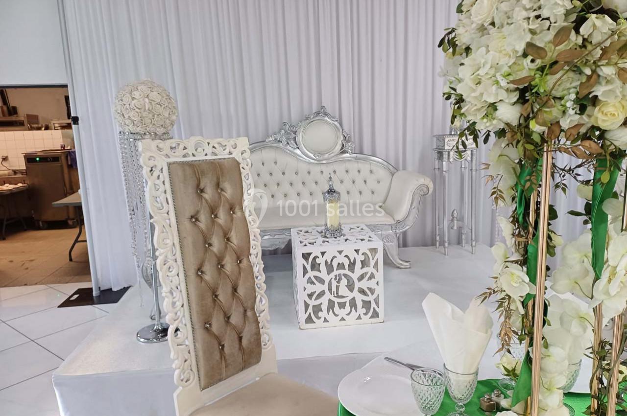 Salle décorée pour un événement avec canapé blanc, table ornée, chaise capitonnée et fleurs blanches en premier plan.