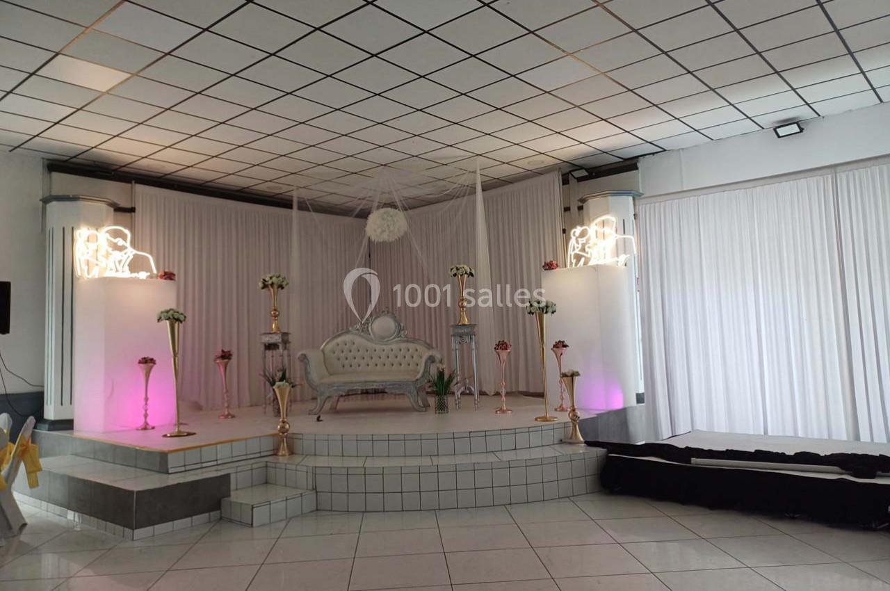 Scène de mariage décorée avec un canapé blanc, colonnes lumineuses, fleurs et rideaux blancs en arrière-plan.