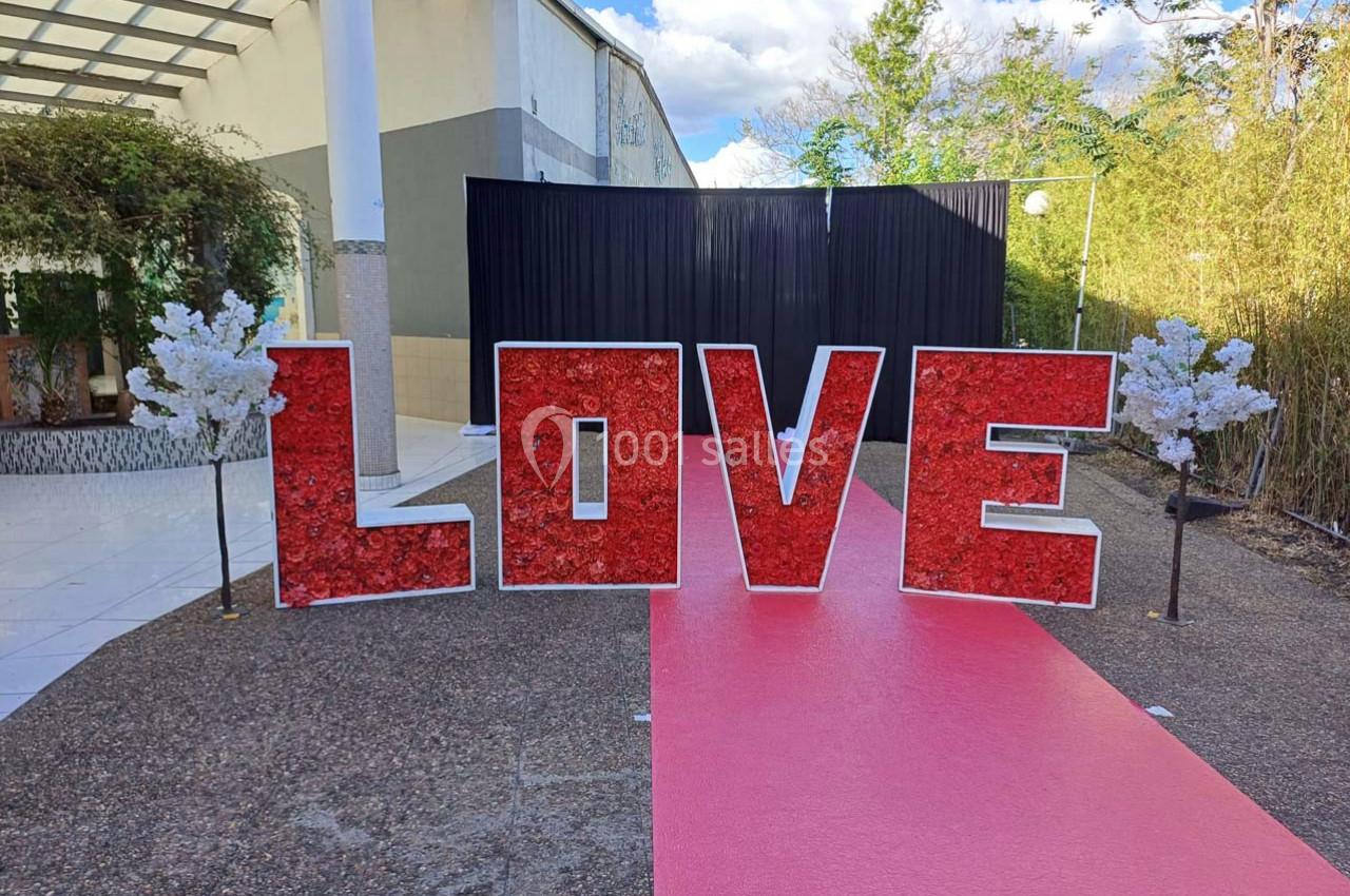 Lettres rouges formant le mot ’LOVE’ sur un tapis rouge, entourées de petites décorations blanches, en extérieur.