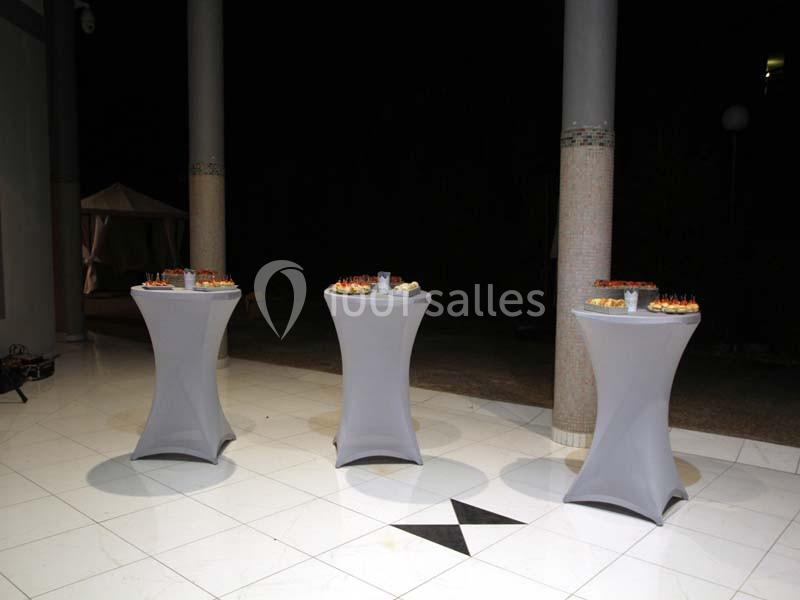 Trois tables hautes recouvertes de nappes blanches, disposées sur un sol carrelé, avec des amuse-bouches dessus.