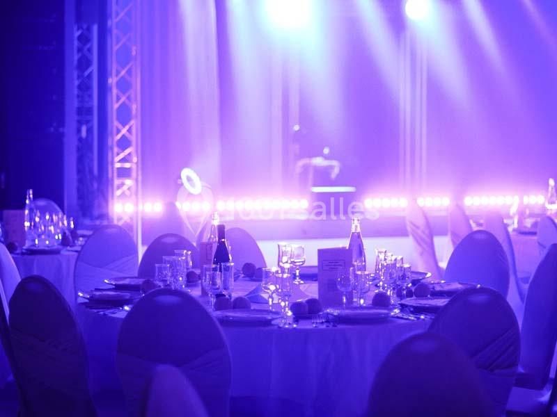 Salle de réception éclairée par des lumières violettes, avec des tables rondes dressées pour un événement.