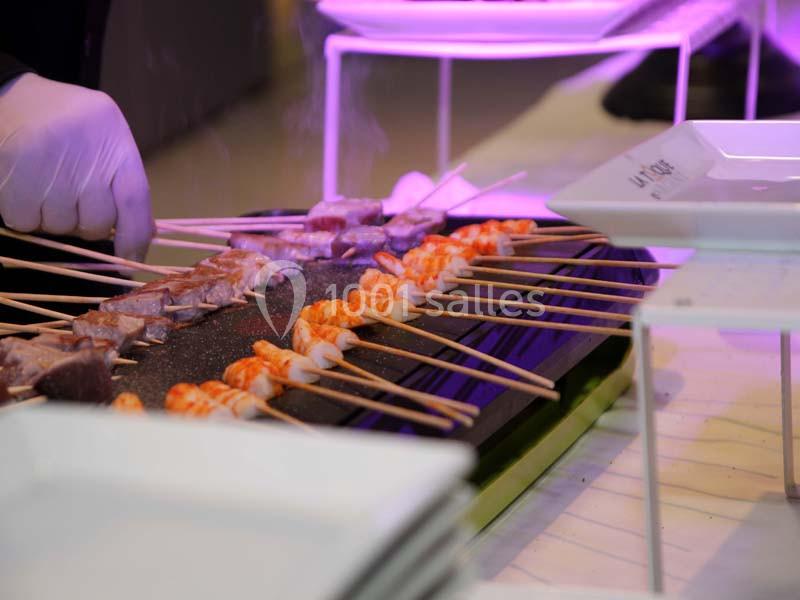 Brochettes de crevettes et de viande grillées sur une plaque chauffante, préparées par une personne portant des gants.