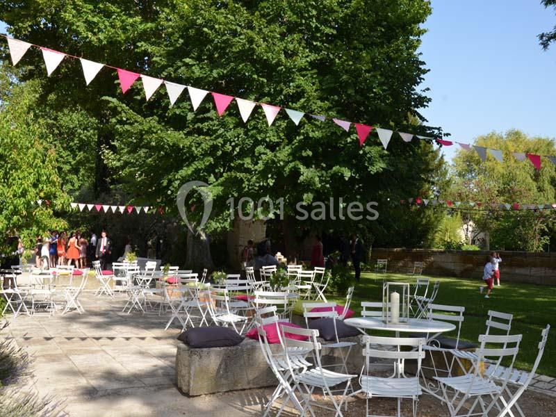 Chaises et tables blanches disposées en extérieur sous des guirlandes colorées, dans un jardin arboré par temps ensoleillé.