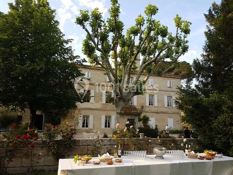 Table dressée avec des boissons et amuse-bouches devant une grande maison en pierre entourée de jardins fleuris.