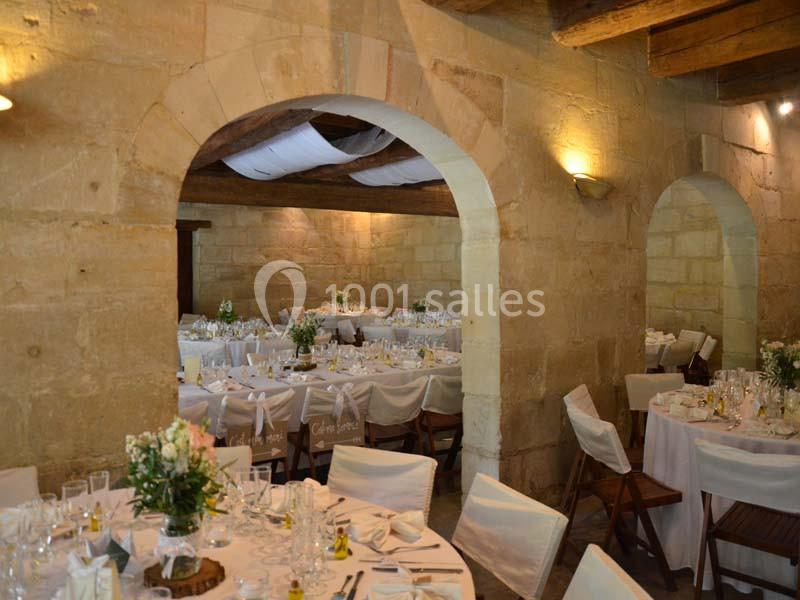 Salle en pierre avec tables rondes dressées pour un événement, décorées de nappes blanches et de fleurs.