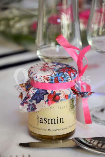 Petit pot de miel au jasmin décoré d’un tissu fleuri et d’un ruban rose, posé sur une table dressée.