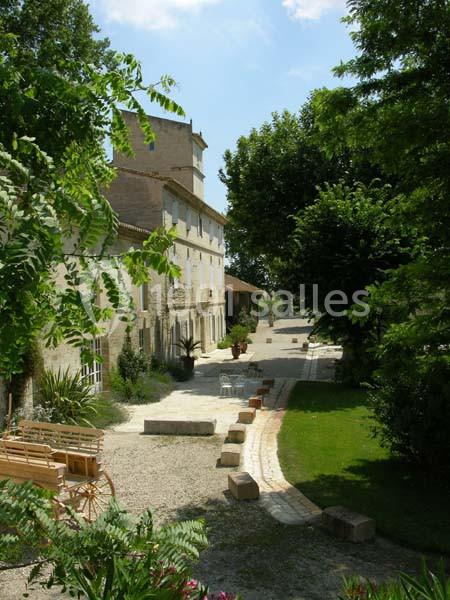 Vue d'une bâtisse en pierre entourée de verdure, avec une allée pavée et des bancs en bois dans un jardin.