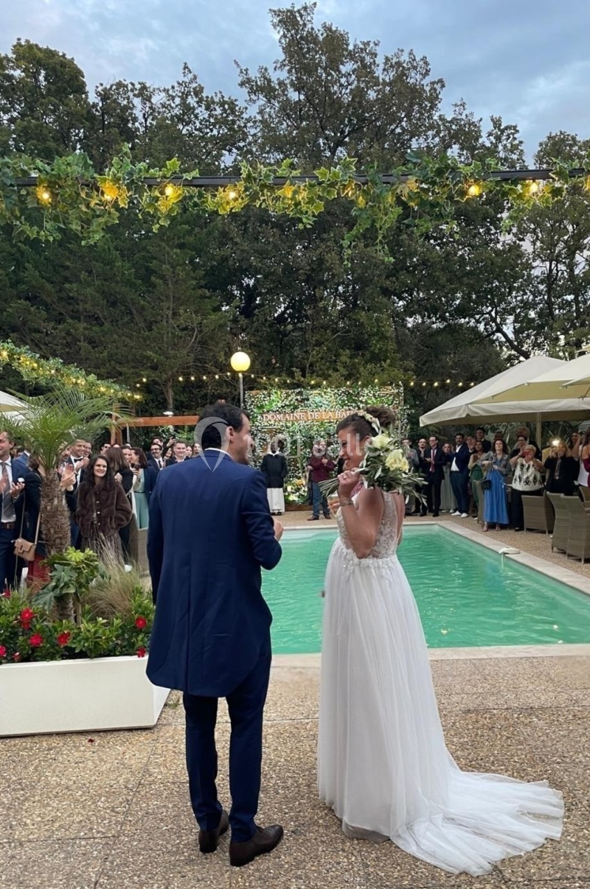 Un couple en tenue de mariage échange devant une piscine, entouré d'invités et de décorations lumineuses extérieures.
