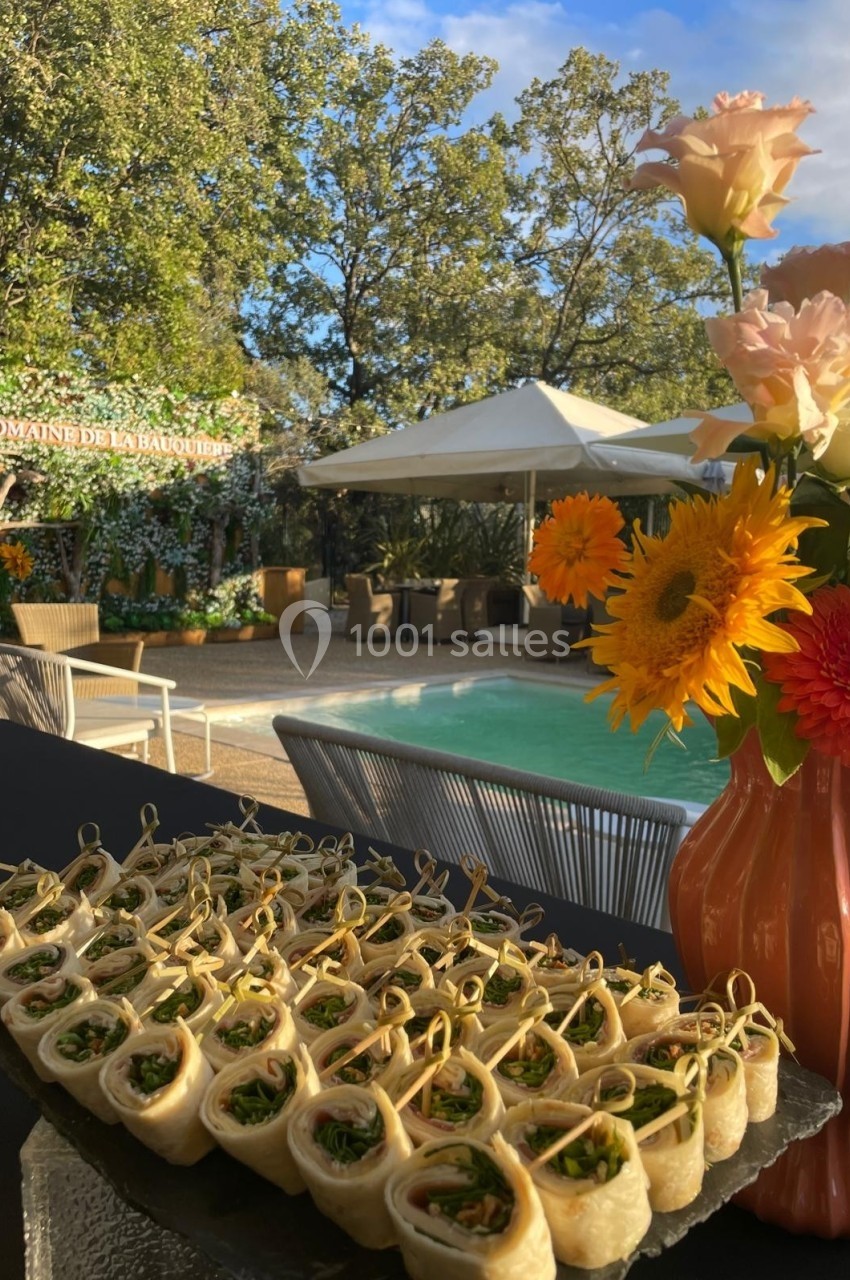 Plateau de bouchées roulées garnies, disposé près d'une piscine entourée de fleurs et de mobilier de jardin.