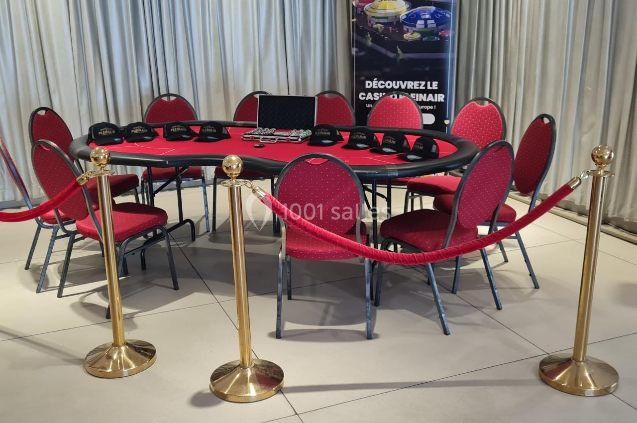 Table de poker rouge entourée de chaises, cordons de sécurité dorés et affiches en arrière-plan.