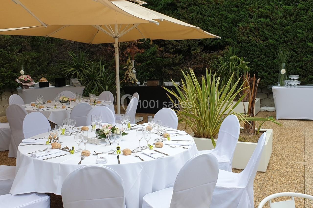 Tables rondes dressées avec nappes blanches et décorations florales, disposées en extérieur sous des parasols.