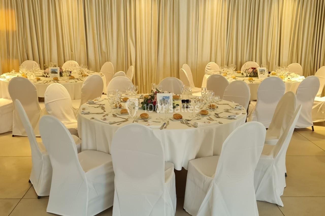 Salle de réception avec des tables rondes dressées, nappes blanches, chaises couvertes et décorations de table.