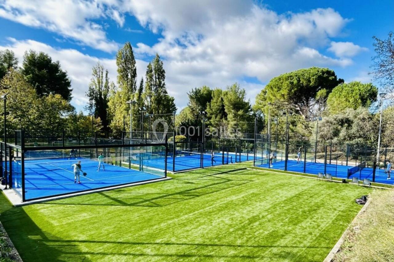 Terrains de padel extérieurs avec gazon synthétique, entourés d'arbres et sous un ciel partiellement nuageux.